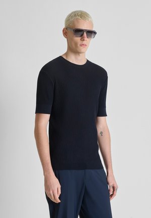 Uomo con capelli biondi corti, che indossa una maglietta nera a coste a maniche corte, pantaloni blu navy, grandi occhiali da sole neri e un piccolo tatuaggio sull'avambraccio sinistro.