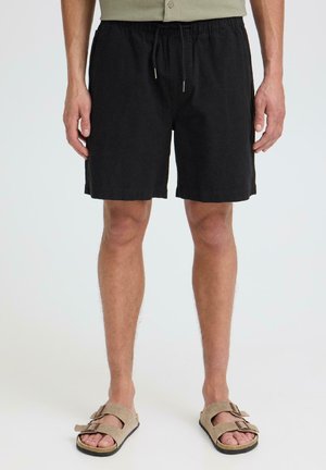 Homme portant un short noir à cordon de serrage et des sandales en daim beige, debout devant un fond clair uni.