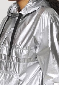 Silver vattentät jacka med en blank yta, justerbar huva och svarta dragsnören. Har en framficka och lättviktig design.