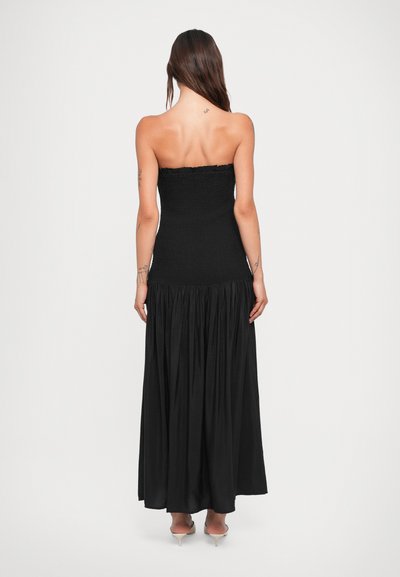 Rochie maxi neagră, fără bretele, cu un corset elastic și fustă plisată. Textură netedă, până la podea, cu tocuri transparente înalte până la glezne vizibile.