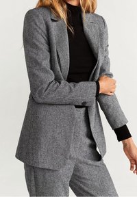 Blazer gris sur mesure en tissu texturé, avec larges revers, fermeture par un seul bouton et poches en biais. Porté sur un col roulé noir côtelé.