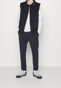 Marineblauwe mouwloze vest met knopen aan de voorkant over een lichtgrijze lange mouwen shirt, gecombineerd met marineblauwe tapered broek en grijze sneakers met blauwe accenten.