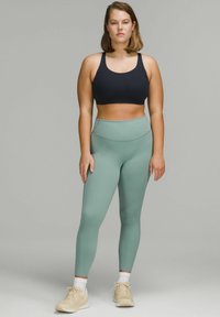 Zwarte sportbeha met brede banden, gecombineerd met high-waisted mintgroene leggings met een gladde textuur. Model draagt beige sneakers.