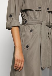 Trench coat color khaki con bottoni neri, cintura e maniche arrotolate; texture liscia; include tasche frontali e un design del colletto classico.