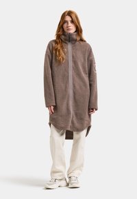 Brun fleece-jakke med høj krave og frontlynlås, med lange ærmer og en længere ryg for ekstra dækning. Hvide bukser og sneakers synlige.