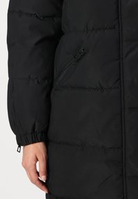 Svart pufferjacka med slät textur, kviltat mönster, elastiska ärmmuddar och en zip-ficka på sidan.