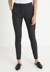 Žena ve tmavě šedých kalhotech s tenkými pruhy, bílém topu a černých špičatých botách bez tkaniček, stojící s jednou rukou v kapse.