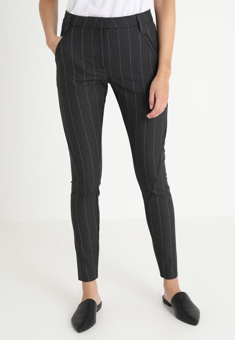 Žena ve tmavě šedých kalhotech s tenkými pruhy, bílém topu a černých špičatých botách bez tkaniček, stojící s jednou rukou v kapse.