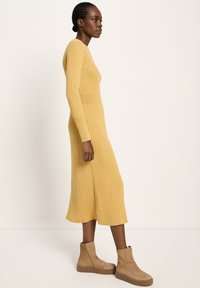 Frau in einem senfgelben, gerippten Midikleid mit langen Ärmeln, kombiniert mit beigefarbenen, plateauförmigen Stiefeletten, vor einem beigen Hintergrund stehend.