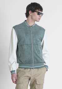 Giacca bomber in suede verde con maniche bianche, polsini a coste e chiusura con zip. Presenta un sottile dettaglio del logo sul petto.