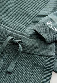 Groene ribbeltrui met een subtiele visgraatpatroon. Voorzien van een trekkoord in de taille en een logo op de manchet. Zachte textuur.
