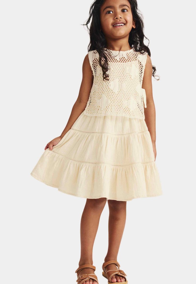 Niña joven sonriente mientras sostiene el dobladillo de su vestido sin mangas de color crema con parte superior de ganchillo y falda de capas, llevando sandalias color cuero.