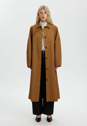Femme blonde portant un long manteau marron boutonné par-dessus un col roulé blanc et un pantalon noir à jambes larges, debout devant un fond clair.