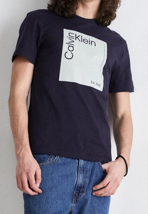Person wearing a navy blue Calvin Klein t-shirt with a large white square logo and blue jeans, one hand in pocket and bracelet visible.