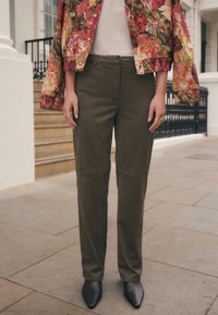 Pantalon sur mesure vert olive avec une texture lisse et des détails de couture, associé à des chaussures noires à bout pointu et une veste à motif floral.