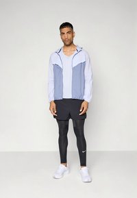 Lichtgewicht jas met ritssluiting in lichtblauw en wit, gecombineerd met zwarte sportshorts en reflecterende leggings. Witte sportschoenen maken de look compleet.