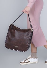 Bruine leren tote bag met studaccenten, ritssluiting en twee schouderbanden. Gladde textuur; tas is groot en slungelig van vorm.