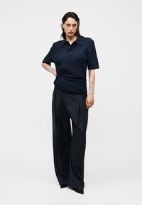 Νεαρό άτομο με τατουάζ που φοράει μαύρο πουκάμισο με κοντά μανίκια και γιακά και φαρδιά μαύρα παντελόνια, στέκεται με το ένα χέρι στη μέση.