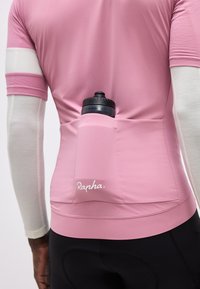 Egy kerékpáros rózsaszín Rapha mezben, hátul zsebben vízpalackkal, fehér karujjakkal és fekete kerékpáros rövidnadrágban.