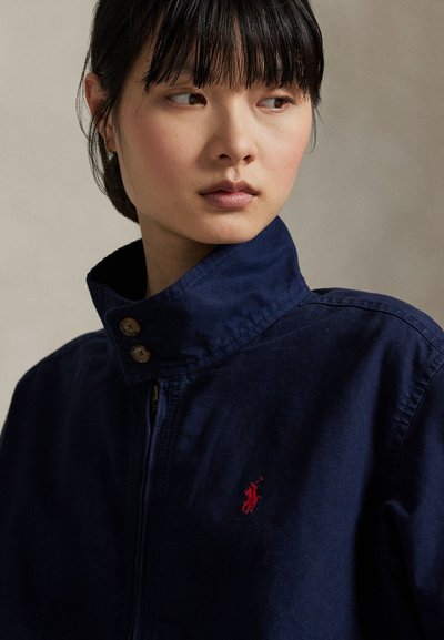 Ung kvinde med mørkt hår og pandehår iført en mørkeblå jakke med opretstående krave og en lille rød broderi på brystet.