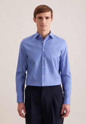 Giovane uomo con camicia azzurra a maniche lunghe con motivo a bottoni e pantaloni neri, in piedi contro uno sfondo neutro semplice.