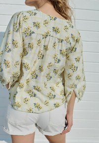 Bloemdrukblouse met gele bloemen en groene accenten op een witte geblokte achtergrond. Losse fit, driekwartmouwen, gestructureerde stof.