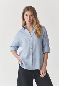 Camisa de botones azul claro hecha de tejido texturizado, con mangas enrollables y botones de madera, acompañada de pantalones negros.