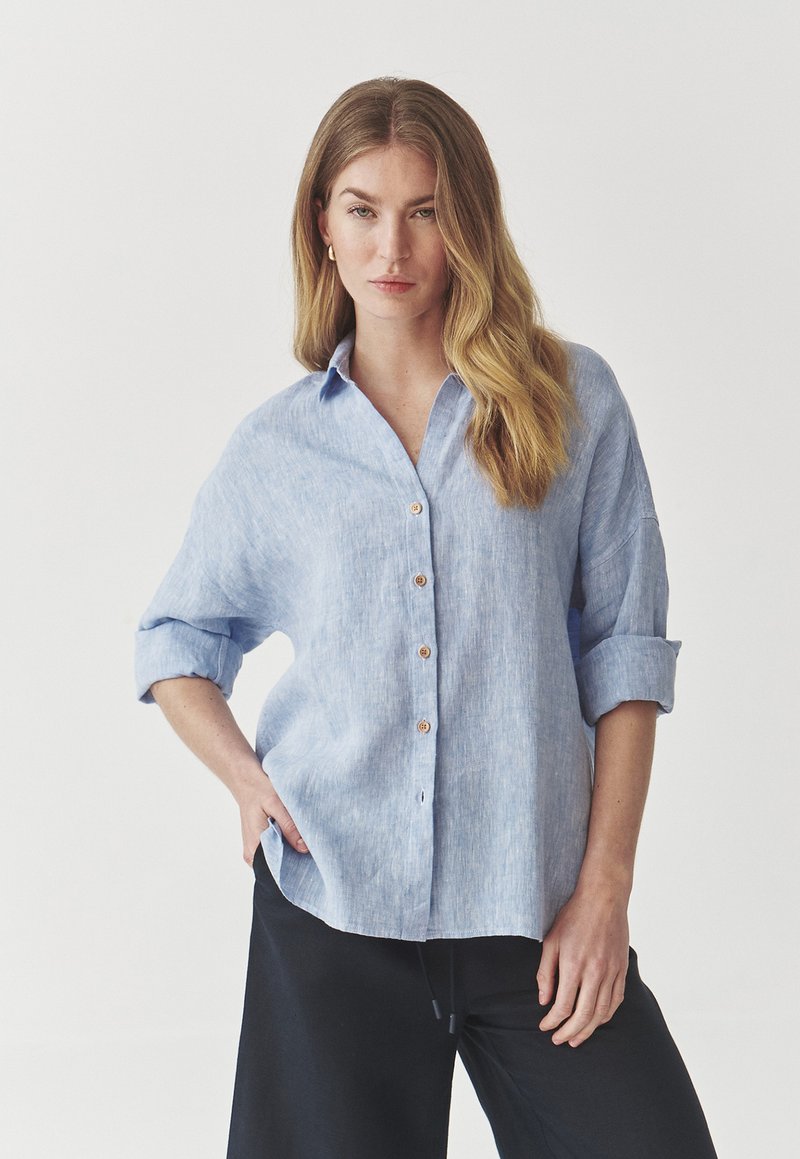 Camisa de botones azul claro hecha de tejido texturizado, con mangas enrollables y botones de madera, acompañada de pantalones negros.