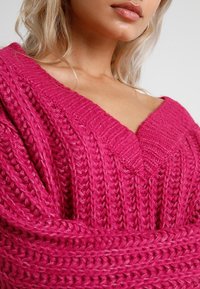 Pull rose tricoté avec un col en V, un motif texturé et des manches larges. Le tissu semble doux et épais, idéal pour superposer.