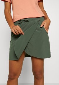 Falda envolvente verde oliva con una textura suave, que presenta una cintura con cordón y bolsillos laterales. El dobladillo cae a mitad de muslo con un diseño en capas.