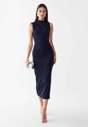 Femme debout dans une robe midi bleu marine sans manches ajustée, tenant une pochette argentée, portant des talons et des boucles d'oreilles argentés, sur fond blanc.