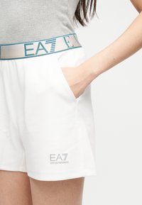 Femme portant un short blanc EA7 Emporio Armani avec le logo sur la jambe gauche et la main dans la poche, assorti à un haut gris sans manches.