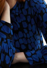 Blue and black patterned blouse featuring an abstract design with shapes in various sizes. Long sleeves with button accents and gathered details.