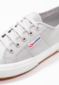 Baskets en toile grise avec des lacets gris clair, des œillets en métal, une semelle en caoutchouc blanche et un petit logo rouge et bleu sur le côté.