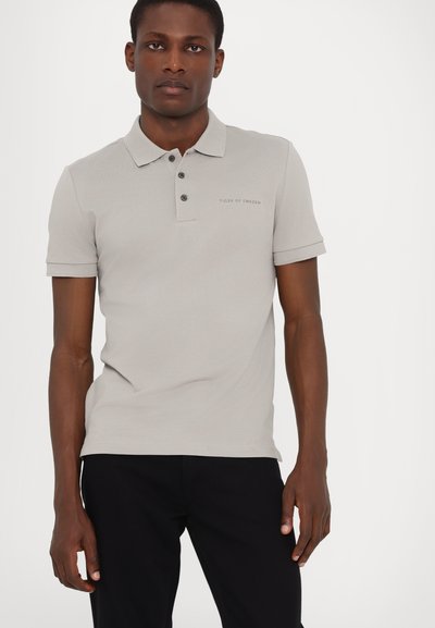 Helehall polo-särk, mis on valmistatud tekstureeritud kangast, millel on kaeluse lõige, kolme nupuga nööpide rida ja tikand logoga rinnal.