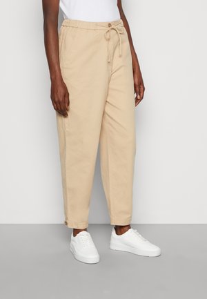Beige, lichte katoenen broek met een elastische tailleband en een trekkoord, voorzien van omslagmanchetten en zijzakken. Gepaard met witte sneakers.