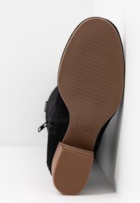 Black suede boot with a tan rubber sole, featuring a smooth texture and horizontal grooves for grip. Branding is visible on the sole.