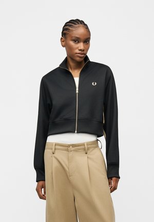 Nuori nainen, jolla on letitetty hiukset, pukeutuneena mustaan vetoketjulliseen takkiin valkoisen paidan ja beigenväristen korkeavyötäröisten housujen päällä, seisoo vaaleaa taustaa vasten.