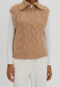 Gilet sans manches en maille torsadée couleur fauve avec col demi-fermeture éclair, porté sur un haut blanc à manches longues et associé à un pantalon blanc taille haute.