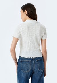 Camisa blanca de textura y manga corta con cuello, combinada con vaqueros azules de cintura alta. Enfócate en el patrón ondulado de la camisa y su diseño recortado.