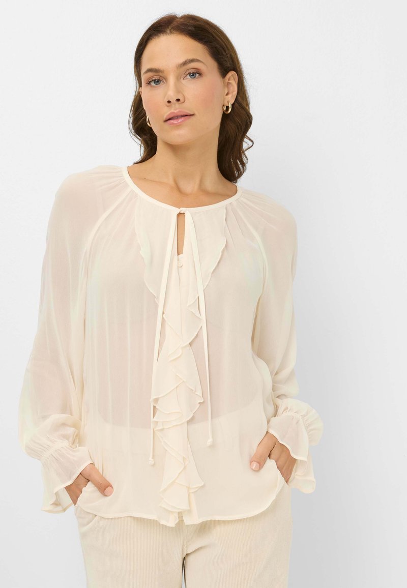 Cream blouse with ruffled front, round neckline, and drawstring closure. Long sleeves with gathered cuffs. Lightweight, sheer fabric.