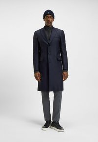Uomo in un lungo cappotto blu navy, pantaloni grigi, sneaker nere, maglione scuro con zip e berretto blu navy, in piedi su uno sfondo semplice e chiaro.