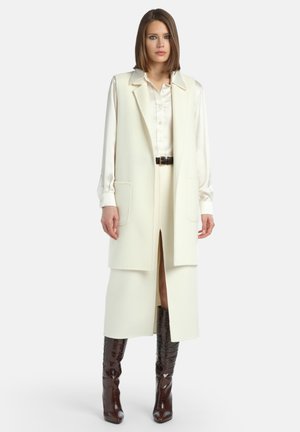 Long gilet créme fabriqué en tissu doux, avec une ouverture sur le devant, un col cranté et deux poches poitrine, associé à un chemisier créme et des bottes marron.