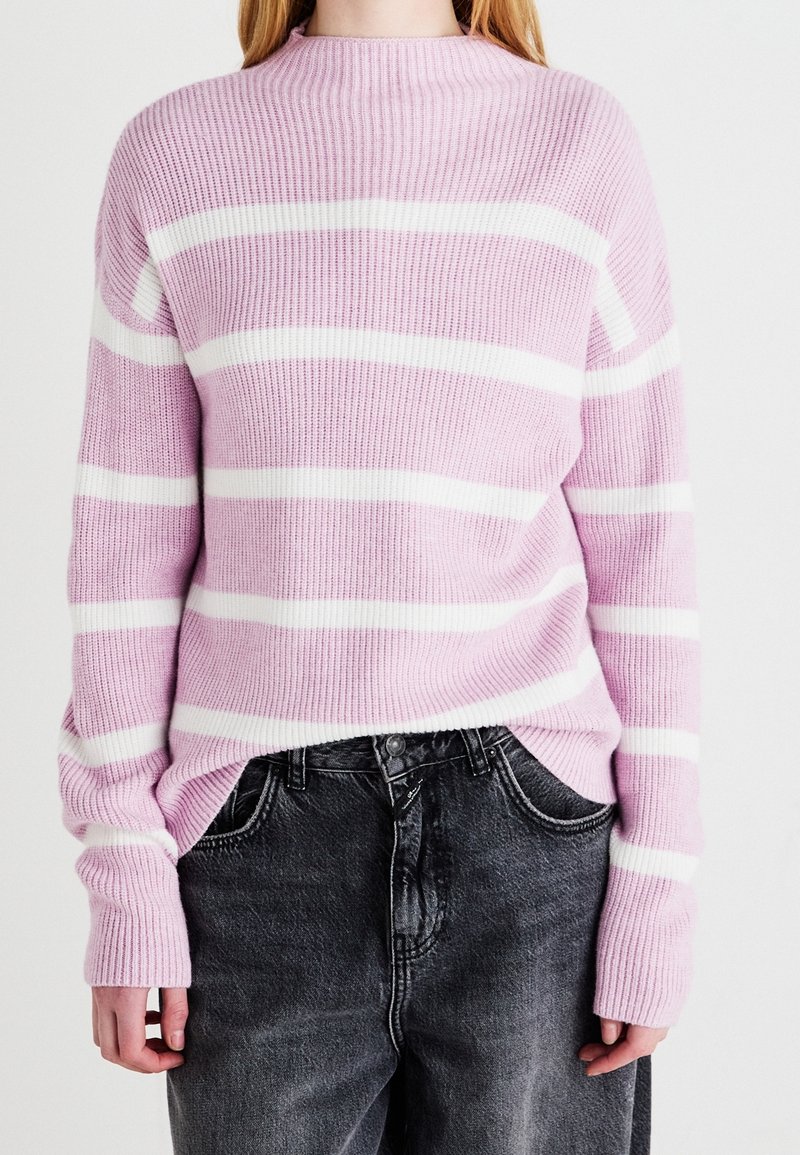 Różowy i biały sweter w paski dziany o prążkowanej fakturze, z okrągłym dekoltem i lekko za dużymi rękawami, zestawiony z ciemnymi jeansami o wyraźnej fakturze.
