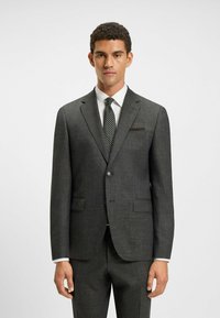 Abito grigio scuro in tessuto testurizzato, chiusura con bottone singolo, revers a lancia e fazzoletto da taschino. Completato con una camicia bianca e una cravatta a pois.