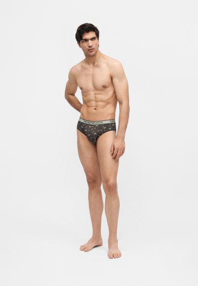 Μυώδης άνδρας στέκεται ξυπόλητος, φοράει σκούρα σλιπ με σχέδια και ζώνη "EMPORIO ARMANI", πόζαρε σε απλό φωτεινό φόντο.