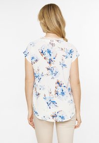 Blumenbluse mit cremefarbenem Hintergrund, verziert mit blauen und braunen Blumen. Kurze Ärmel, runder Ausschnitt und Spitzenverzierung am Kragen.