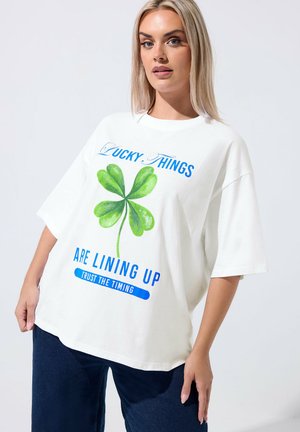 Blondynka w białej, oversize'owej koszulce z dużym zielonym koniczyną czterolistną i niebieskim napisem o szczęściu i czasie.