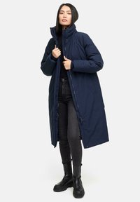Lang navy blå pufferjakke med høj krave, lynlås lukning og sidelommer. Båret over en sort top og parret med sorte jeans og støvler.