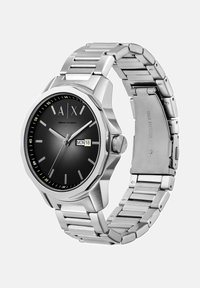 Kovinska ura iz nerjavnega jekla z okroglo črno številčnico, srebrnimi oznakami ur in prikazom datuma. Ima srebrn kovinski pas in osemkotno ohišje.